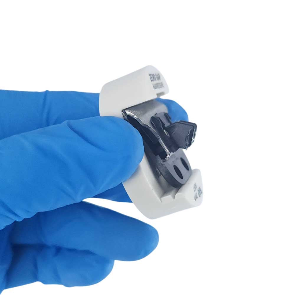 A hand wearing a blue nitrile glove holds a small white plastic component with a black and silver metal clip or connector against a plain white background.